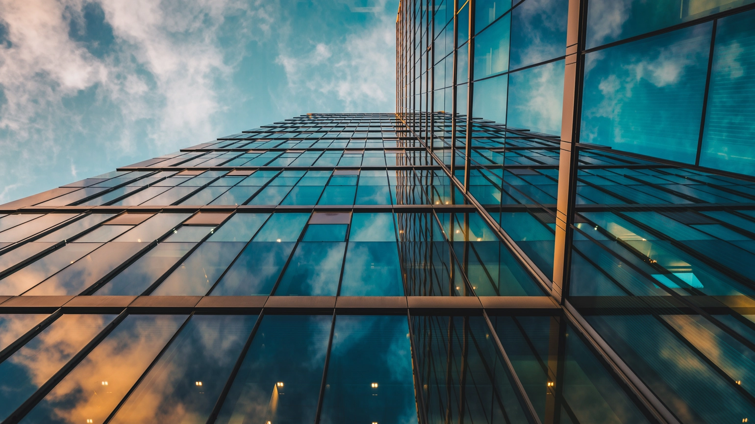 Upward view of modern glass skyscraper reflecting blue sky and clouds, with geometric lines and warm interior lights at dusk. Upward view of modern glass skyscraper reflecting blue sky and clouds, with geometric lines and warm interior lights at dusk.